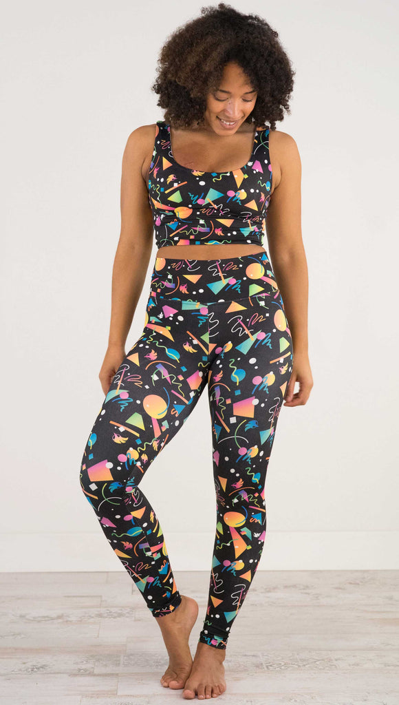 Full Body view of model wearing WERKSHOP white confetti athleisure leggings. The artwork on the leggings shows multi-colored circles, scribbles and triangles over a black background.