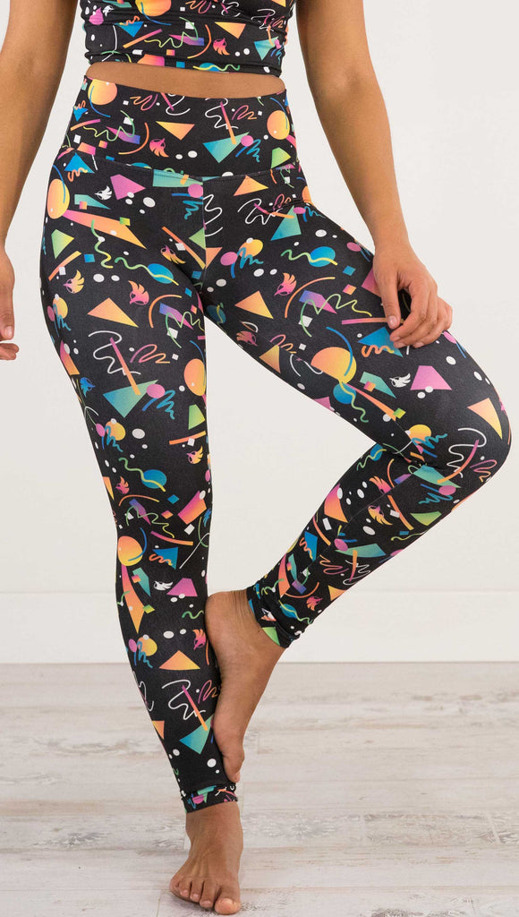 Waist down view of model wearing WERKSHOP white confetti athleisure leggings. The artwork on the leggings shows multi-colored circles, scribbles and triangles over a black background.