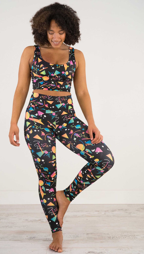 Full Body view of model wearing WERKSHOP white confetti athleisure leggings. The artwork on the leggings shows multi-colored circles, scribbles and triangles over a black background.