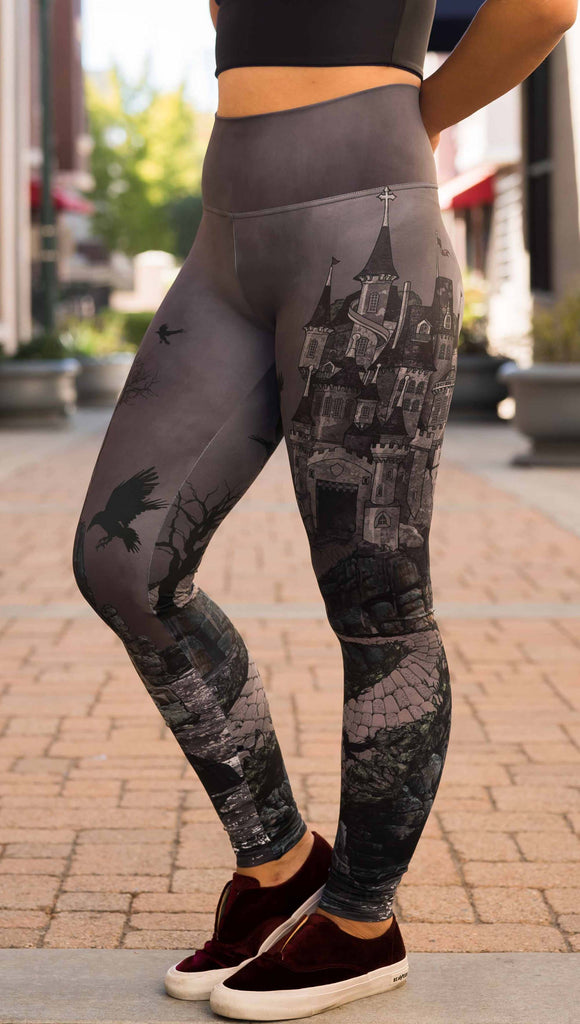 Model wearing WERKSHOP Dark Castle Leggings. The artwork on the leggings feature a fully monochrome (black and gray) gothic castle with stone walkway and crows flying in the moonlight.