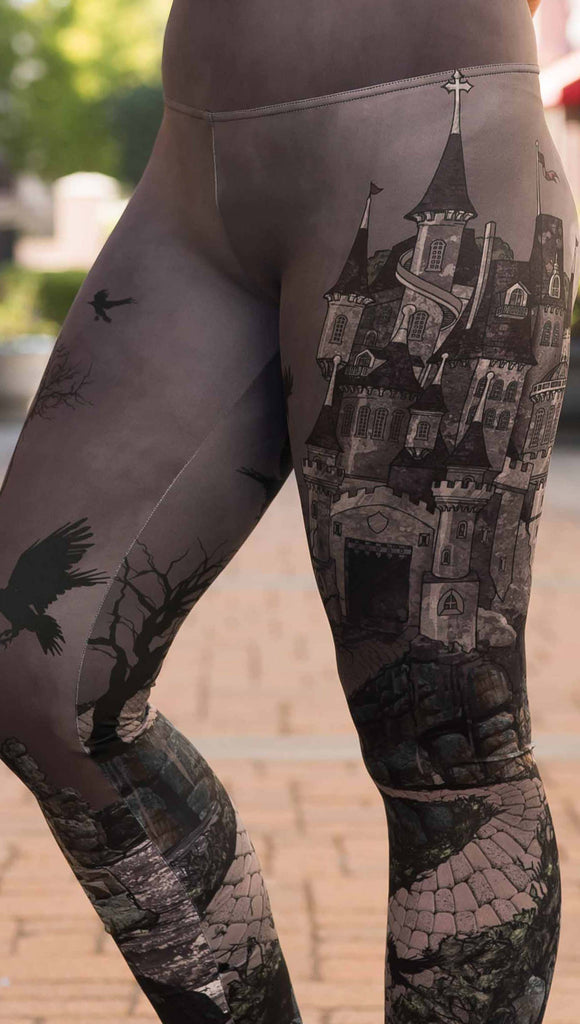 Zoomed in view of model wearing WERKSHOP Dark Castle Leggings. The artwork on the leggings feature a fully monochrome (black and gray) gothic castle with stone walkway and crows flying in the moonlight.