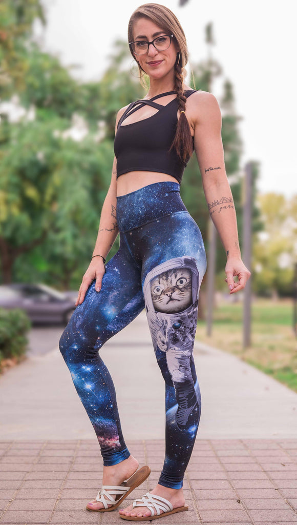 Model wearing Catstronaut Leggings. The leggings are printed with a cat in an astronaut suit on the wearer's left leg. The galaxy background is a deep cobalt blue with nebula swirls and shooting stars.