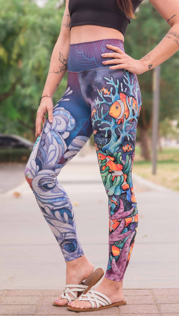 Model wearing WERKSHOP Clownfish and Surf Mashup Leggings. These leggings feature two iconic prints. on the right leg, you have a surfboard with dolphins and spashing waves. On the left leg, you have an explosion of color with a coral reef and clownfish.