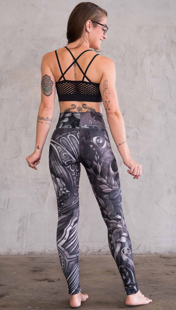 Zoomed out back view of model wearing the gargoyle mashup leggings in a black and gray color. One leg has a gargoyle and the other leg has a large skull