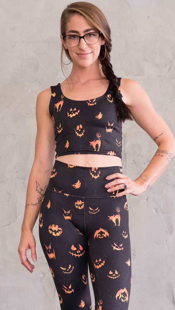 Front view of model wearing the reversible Boo! and Jack-O-Lantern crop top. This is in the Jack-O-Lantern side, it is in black with bright orange jack-o-lantern faces and cats throughout