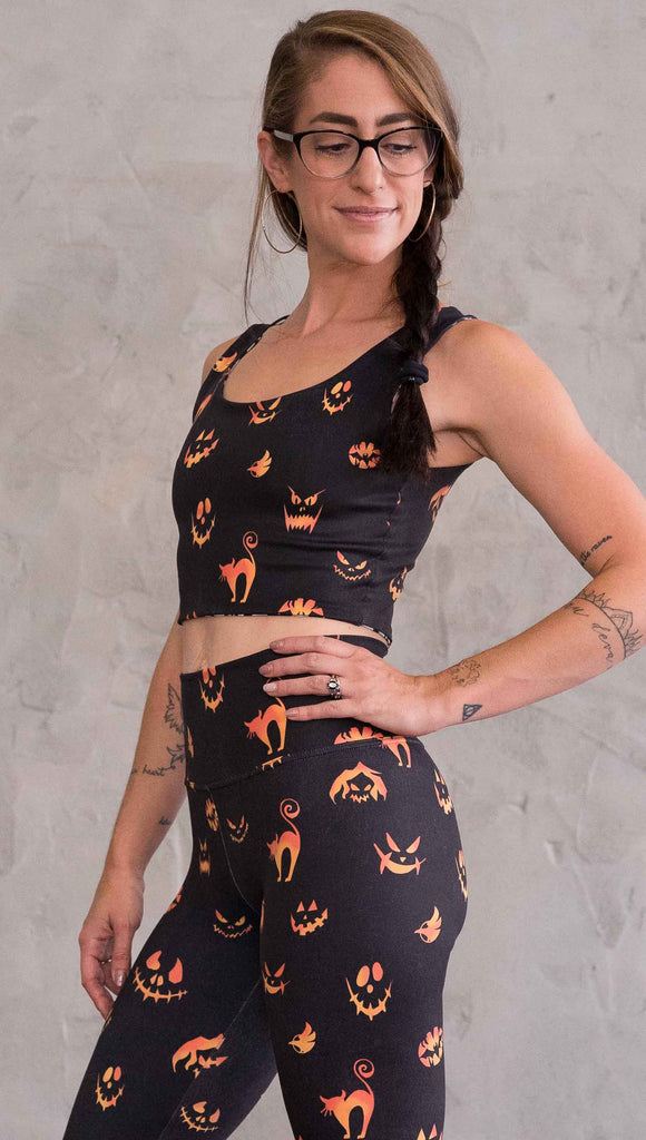 Left view of model wearing the reversible Boo! and Jack-O-Lantern crop top. This is in the Jack-O-Lantern side, it is in a black with bright orange  jack-o-lantern faces and cats throughout