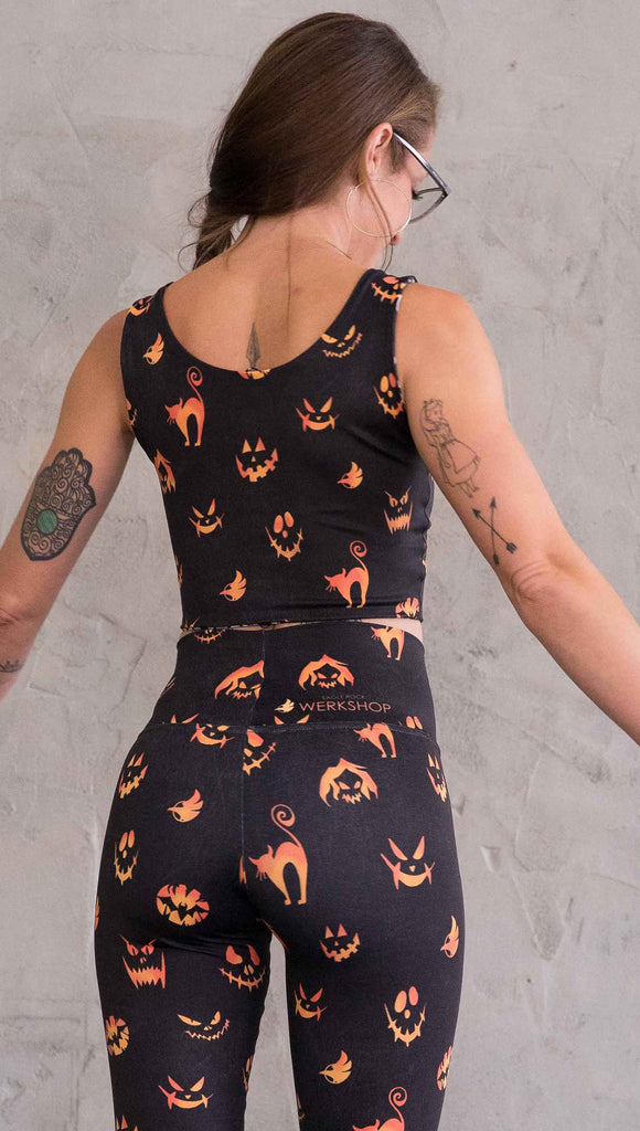 Back view of model wearing the reversible Boo! and Jack-O-Lantern crop top. This is in the Jack-O-Lantern side, it is in black with bright orange jack-o-lantern faces and cats throughout