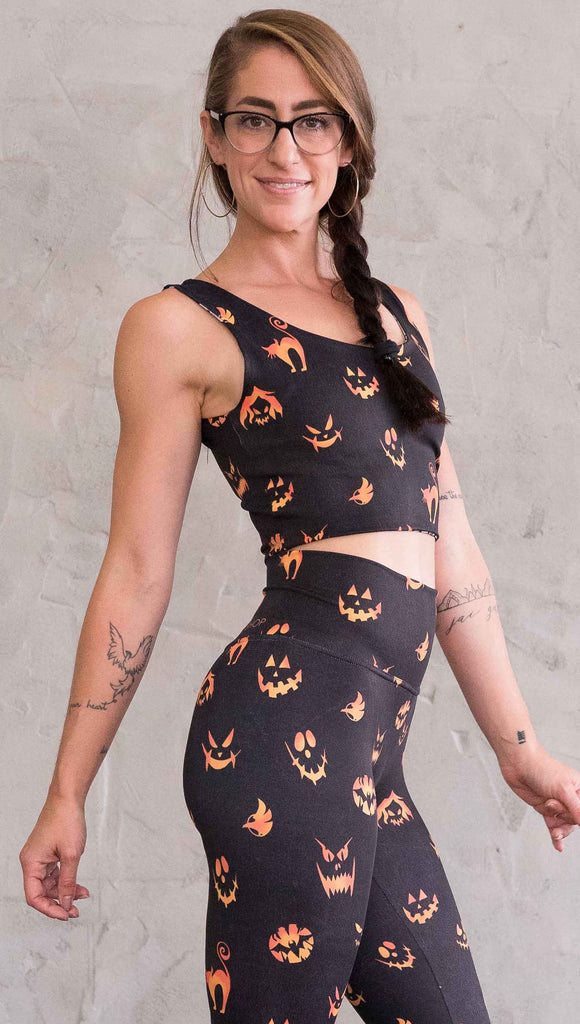 Right  view of model wearing the reversible Boo! and Jack-O-Lantern crop top. This is in the Jack-O-Lantern side, it is in black with bright orange jack-o-lantern faces and cats throughout