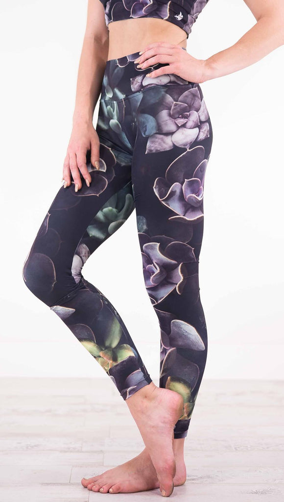 Enhanced left view of model wearing black athleisure leggings with green and purple succulent plants throughout