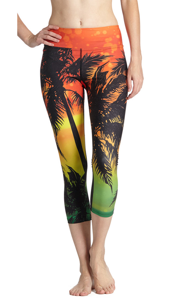 Model wearing WERKSHOP Rasta Palms Leggings. The leggings are printed with large silhouette of palm trees over a rasta themed background with orange at the waistband, yellow at the knee and green at the leg opening.