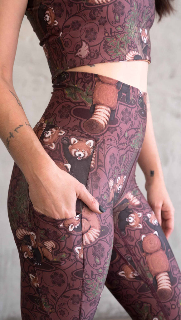 Zoomed in photo of model wearing WERKSHOP Red Panda Leggings. The artwork is dark red with clusters of cute red pandas playing on trees. The leggings have phone pockets on both legs.