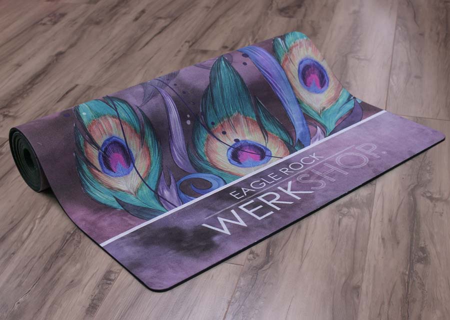Peacock Yoga Mat rolled on the floor with peacock feathers and our logo showing on the bottom edge