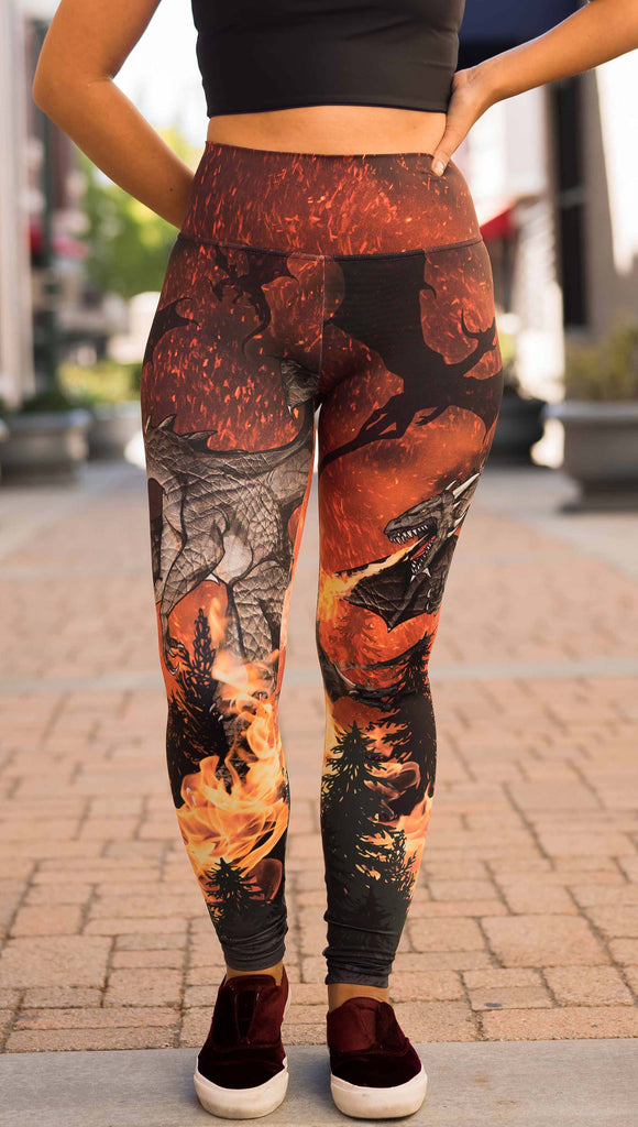 Model wearing WERKSHOP Spitfire Leggings. The artwork on the leggings features a detailed fire breathing dragon blowing flames into a forest with a bright red sky filled with floating embers and silhouettes of more dragons flying in the background.