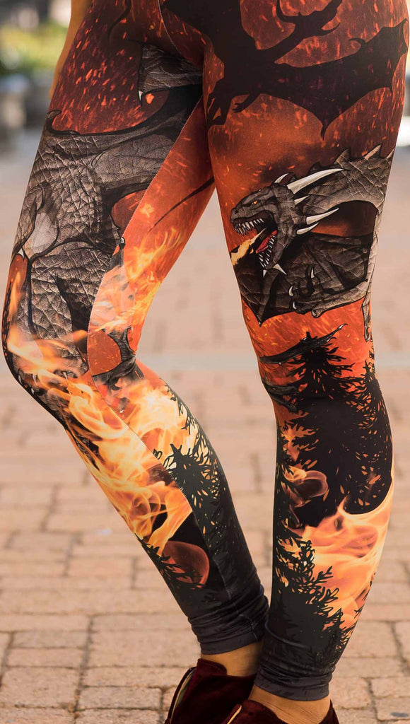 Zoomed in view of model wearing WERKSHOP Spitfire Leggings. The artwork on the leggings features a detailed fire breathing dragon blowing flames into a forest with a bright red sky filled with floating embers and silhouettes of more dragons flying in the background.