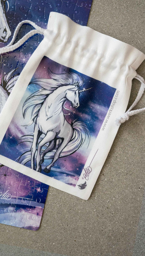 Canvas Drawstring WERKSHOP Unicorn Pouch - comes free with matching 252 piece jigsaw puzzle. The puzzle is printed with original artwork of a Unicorn by Chriztina Marie. It features a white unicorn prancing on a fantastical galactic background with swirls of purple and blue with sparkles.