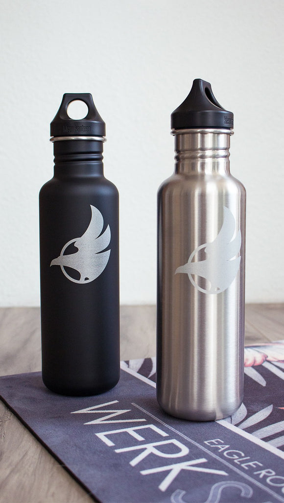 Black water bottle with Eagle Rock Werkshop logo next to silver water bottle with Eagle Rock Werkshop logo resting on a purple yoga mat with Eagle Rock Werkshop logo.