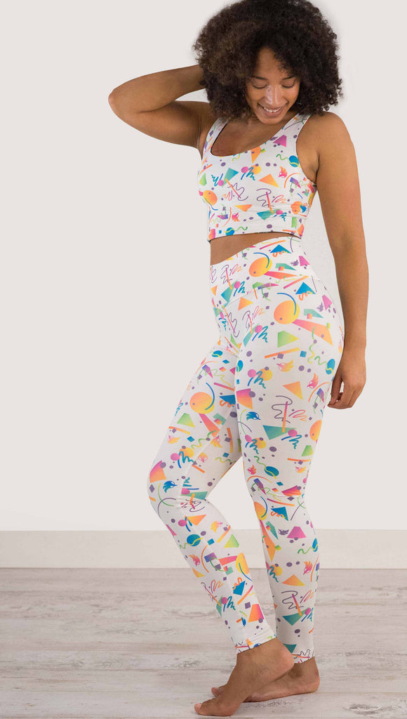 Full body view of model wearing WERKSHOP white confetti athleisure leggings. The artwork on the leggings shows multi-colored circles, scribbles and triangles over a white background.