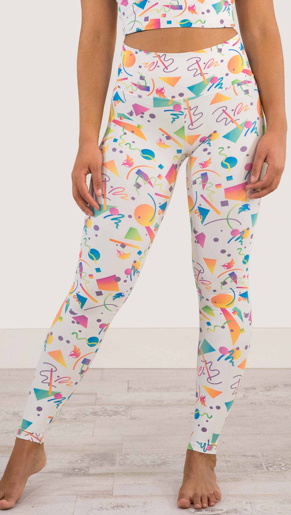 Waist down view of model wearing WERKSHOP white confetti athleisure leggings. The artwork on the leggings shows multi-colored circles, scribbles and triangles over a white background.