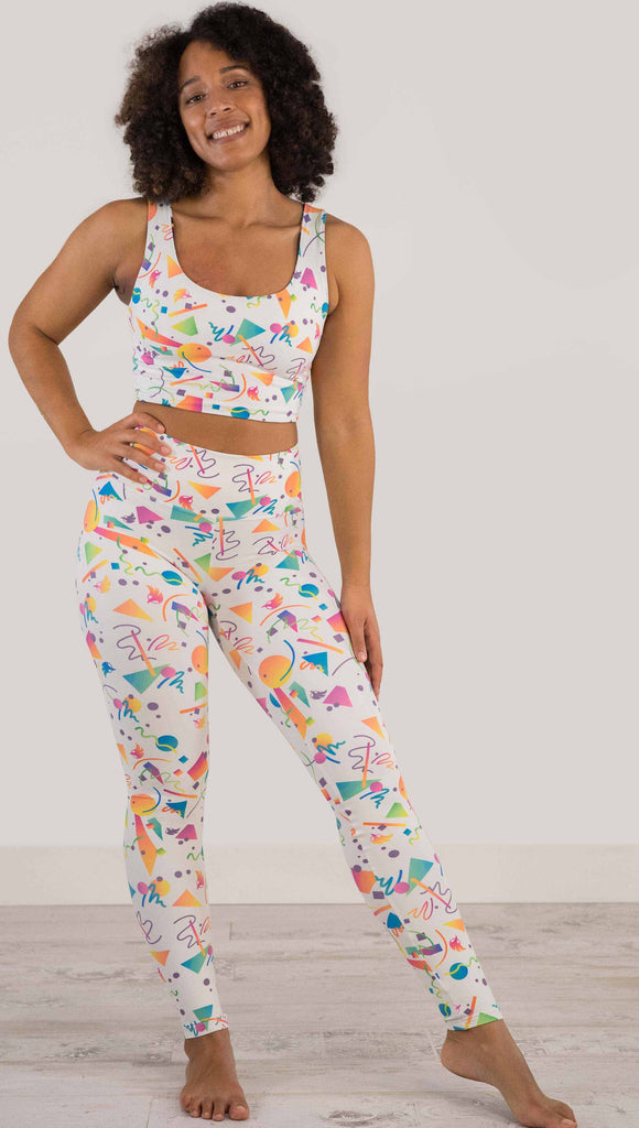 Full body view of model wearing WERKSHOP white confetti athleisure leggings. The artwork on the leggings shows multi-colored circles, scribbles and triangles over a white background.